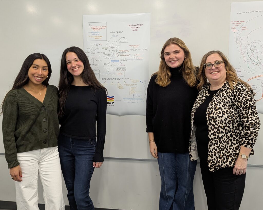 Four women stand in front of a white board with a poster on it.